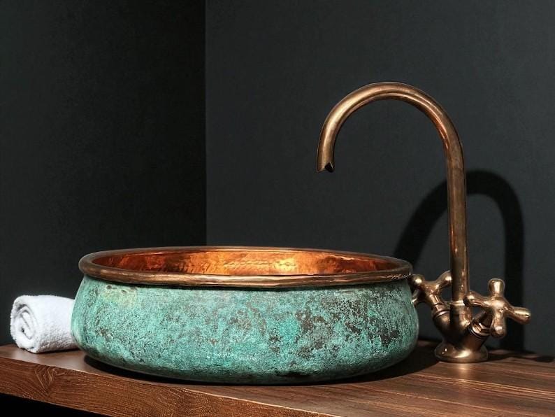 May include: A copper and verdigris patina basin with a bronze faucet. The basin has a rounded shape with a copper rim. The faucet has a curved spout and cross-shaped handles. A rolled white towel sits to the left.