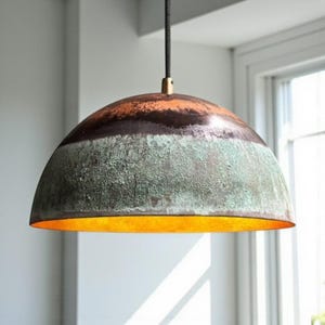 Puede incluir: Lámpara colgante con forma de cúpula y acabado multicolor. La parte superior es marrón y naranja, la central verde y texturizada, y el interior dorado. Se suspende de un cable negro, con un diámetro aproximado de 30 cm.