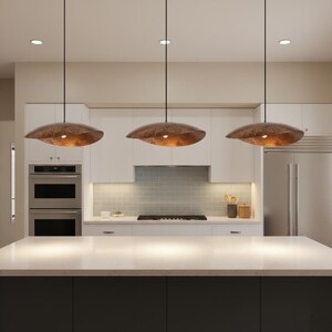 Puede incluir: Tres l&aacute;mparas colgantes de forma org&aacute;nica y color bronce cuelgan sobre una isla de cocina. Las luces emiten un brillo c&aacute;lido. La cocina cuenta con gabinetes blancos y una encimera de color claro.