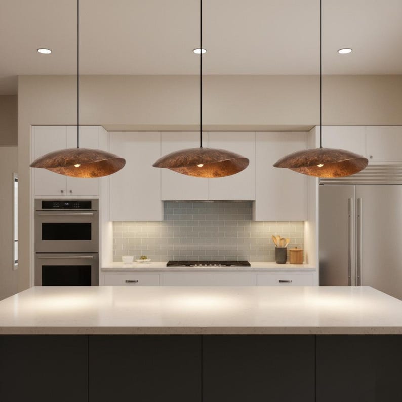 Puede incluir: Tres l&aacute;mparas colgantes de forma org&aacute;nica y color bronce cuelgan sobre una isla de cocina. Las luces emiten un brillo c&aacute;lido. La cocina cuenta con gabinetes blancos y una encimera de color claro.