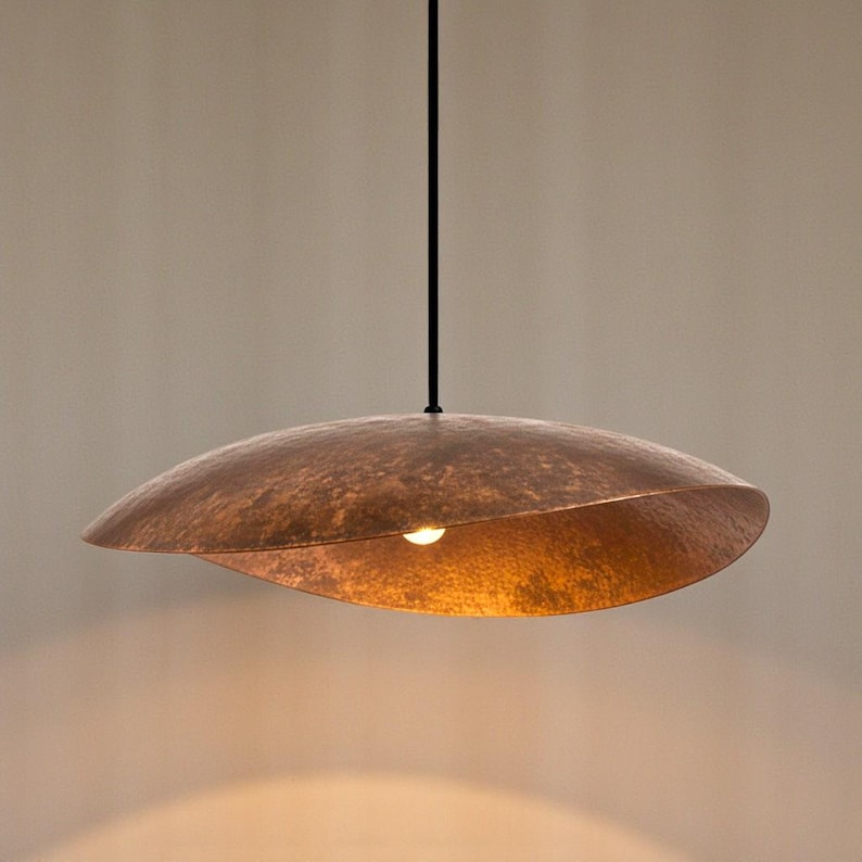 Puede incluir: Una l&aacute;mpara colgante de metal martillado de color cobre. La l&aacute;mpara tiene una pantalla ancha y curvada con una superficie texturizada y una sola bombilla. La l&aacute;mpara cuelga de un cable negro sobre un fondo neutro.