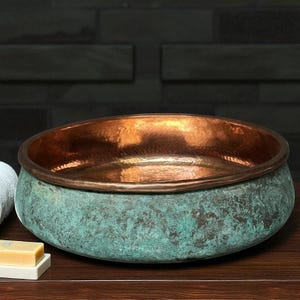 May include: A copper-colored, round sink with a verdigris exterior. The interior of the sink is a shiny copper color. The sink is sitting on a dark wooden surface. The sink is a decorative bathroom fixture.