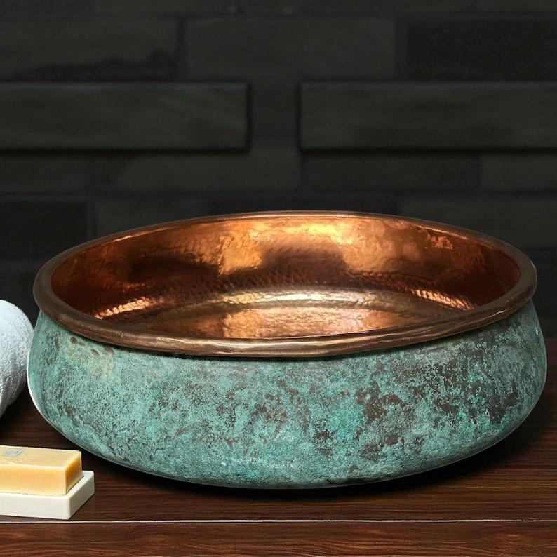 May include: A copper-colored, round sink with a verdigris exterior. The interior of the sink is a shiny copper color. The sink is sitting on a dark wooden surface. The sink is a decorative bathroom fixture.