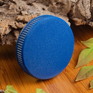 Puede incluir: Un objeto circular azul vibrante con una superficie texturizada y un borde estriado. El objeto est&aacute; sobre una superficie de madera, con un trozo de corteza y hojas verdes en el fondo. El objeto parece ser una tapa o un tap&oacute;n.