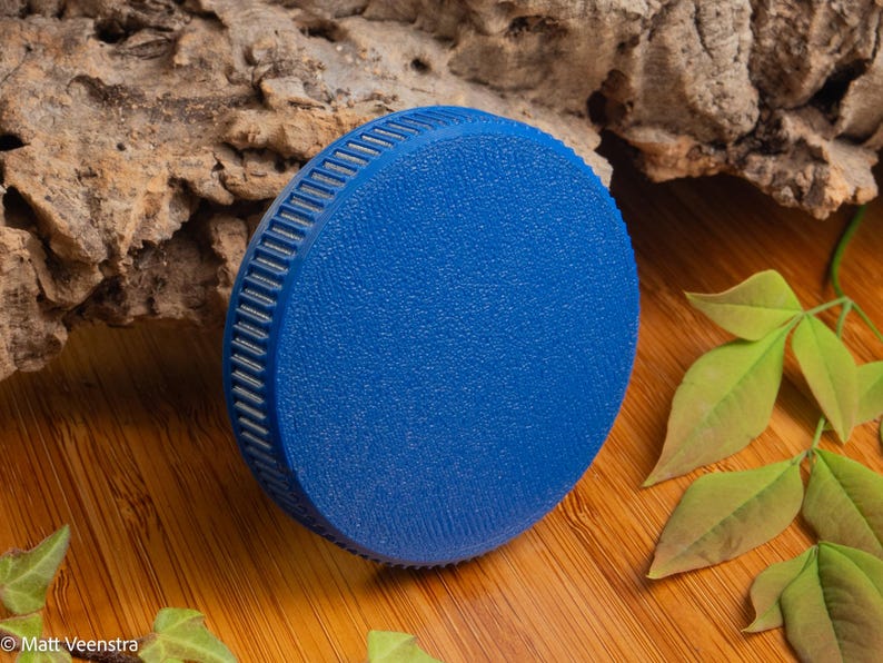 Puede incluir: Un objeto circular azul vibrante con una superficie texturizada y un borde estriado. El objeto est&aacute; sobre una superficie de madera, con un trozo de corteza y hojas verdes en el fondo. El objeto parece ser una tapa o un tap&oacute;n.