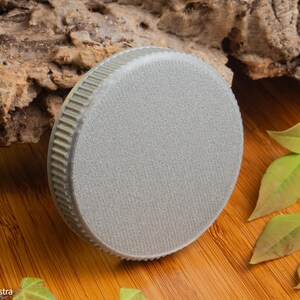 Puede incluir: Un objeto redondo de color gris claro con una superficie texturizada y un borde estriado. El objeto est&aacute; sobre una superficie de madera con hojas verdes y un trozo de madera en el fondo. El objeto parece ser una tapa o un tap&oacute;n.