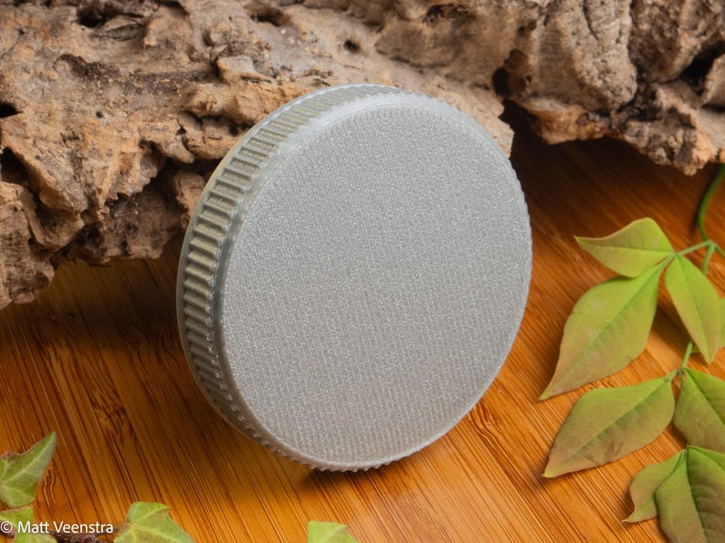 Puede incluir: Un objeto redondo de color gris claro con una superficie texturizada y un borde estriado. El objeto est&aacute; sobre una superficie de madera con hojas verdes y un trozo de madera en el fondo. El objeto parece ser una tapa o un tap&oacute;n.