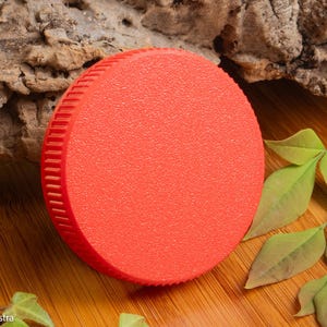Puede incluir: Un objeto circular rojo brillante con una superficie texturizada y un borde estriado. El objeto se encuentra sobre un fondo de madera natural con hojas verdes y un trozo de corteza.