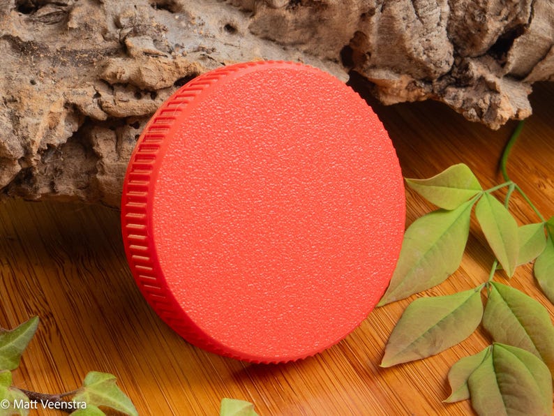 Puede incluir: Un objeto circular rojo brillante con una superficie texturizada y un borde estriado. El objeto se encuentra sobre un fondo de madera natural con hojas verdes y un trozo de corteza.