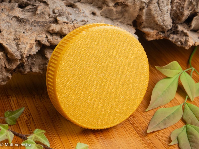 Puede incluir: Un objeto redondo y amarillo con una superficie texturizada y un borde estriado. Se encuentra sobre una superficie de madera, con hojas verdes y corteza en el fondo. El objeto parece ser una tapa o un recipiente.