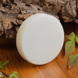 Puede incluir: Una pastilla de jab&oacute;n redonda de color blanquecino con un borde estriado. El jab&oacute;n est&aacute; colocado sobre una superficie de madera con un trozo de corteza y hojas verdes en el fondo. El jab&oacute;n parece no tener perfume y es un producto sencillo y natural.