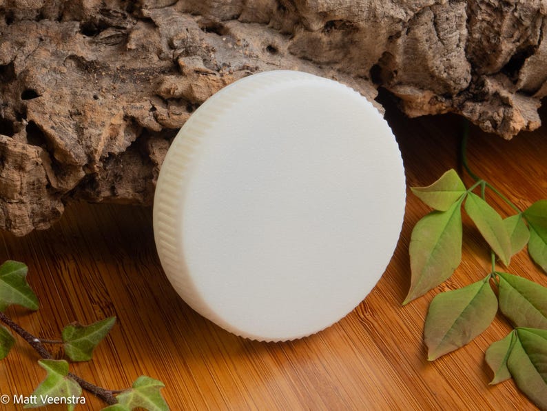 Puede incluir: Una pastilla de jab&oacute;n redonda de color blanquecino con un borde estriado. El jab&oacute;n est&aacute; colocado sobre una superficie de madera con un trozo de corteza y hojas verdes en el fondo. El jab&oacute;n parece no tener perfume y es un producto sencillo y natural.