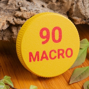 May include: A yellow, circular object with the number "90" and the word "MACRO" printed in red. The object is textured and has a ridged edge. The background includes wood, bark, and green leaves.