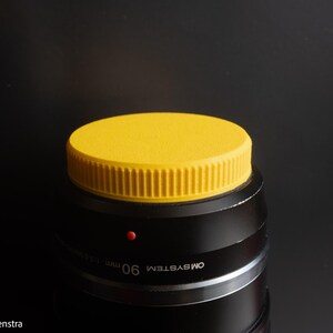 Puede incluir: Un objetivo de c&aacute;mara negro con una tapa de objetivo de color amarillo brillante. La tapa del objetivo es redonda con los lados acanalados. El objetivo tiene el texto "OM SYSTEM" y "90 mm 1:3.5 Macro" impreso. Un peque&ntilde;o punto rojo es visible en el objetivo.