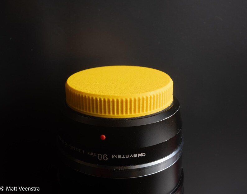 Puede incluir: Un objetivo de c&aacute;mara negro con una tapa de objetivo de color amarillo brillante. La tapa del objetivo es redonda con los lados acanalados. El objetivo tiene el texto "OM SYSTEM" y "90 mm 1:3.5 Macro" impreso. Un peque&ntilde;o punto rojo es visible en el objetivo.