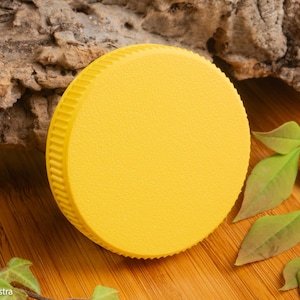 Puede incluir: Una tapa redonda de color amarillo brillante con una superficie texturizada y un borde acanalado. La tapa est&aacute; colocada sobre una superficie de madera con hojas verdes y un trozo de corteza en el fondo. La tapa es de color s&oacute;lido.
