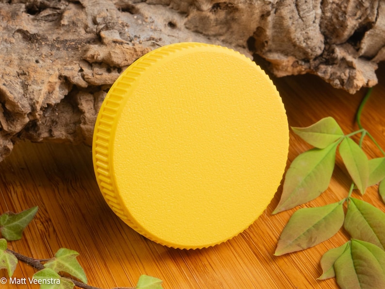 Puede incluir: Una tapa redonda de color amarillo brillante con una superficie texturizada y un borde acanalado. La tapa est&aacute; colocada sobre una superficie de madera con hojas verdes y un trozo de corteza en el fondo. La tapa es de color s&oacute;lido.