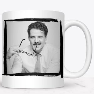 May include: White ceramic mug featuring a black and white image of a man winking and holding glasses. The image is framed by a black border. The mug has a white handle.