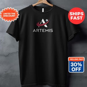 Peut inclure: T-shirt noir avec le mot "ARTEMIS" en blanc et un logo représentant un "A" stylisé avec une ligne incurvée rouge et une sphère blanche. Le t-shirt est suspendu à un cintre en bois. Il y a aussi des autocollants promotionnels.