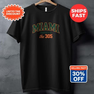 Puede incluir: Camiseta negra con la palabra "MIAMI" en verde y naranja, y "PROPERTY OF the 305" debajo. La camiseta está colgada en una percha de madera sobre un fondo gris.