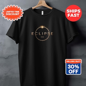 May include: Black t-shirt with a gold eclipse graphic and the word "ECLIPSE" above the year "2024". The shirt hangs on a wooden hanger against a grey background.