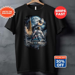 May include: Black t-shirt featuring a detailed graphic of an astronaut on a lunar surface with a rocket launch and planets in the background. The design includes the text "ARTEMIS II".