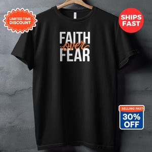 Peut inclure: T-shirt noir avec les mots "FAITH over FEAR" en lettres blanches et orange. Le t-shirt est suspendu à un cintre en bois contre un mur texturé gris. Il y a aussi des autocollants promotionnels.