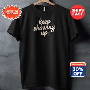 Peut inclure: T-shirt noir avec l'inscription "keep showing up" en écriture cursive. Le t-shirt est suspendu à un cintre en bois contre un mur texturé gris. Il y a des autocollants promotionnels sur le t-shirt.
