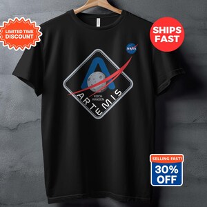 Könnte beinhalten: Schwarzes T-Shirt mit dem Artemis-Missionslogo, einem rautenförmigen Design und einer Mondgrafik. Das NASA-Logo ist ebenfalls sichtbar. Das T-Shirt trägt die Aufschrift "ARTEMIS" und "KOCH HANSEN". Ein Hinweis auf einen Rabatt ist vorhanden.