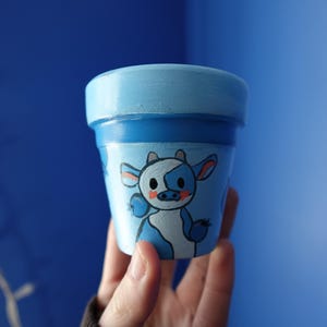May include: A small, hand-painted terracotta plant pot featuring a light blue base color with a darker blue rim. The exterior is decorated with a cartoon-style cow illustration in shades of blue, white, and pink, showcasing a cute character design.