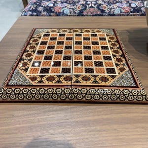 May include: A wooden chess board with a decorative inlaid design. The board features a grid of alternating black and brown squares, with intricate patterns in the borders. The board is set on a wooden table.