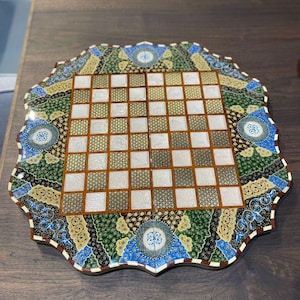 May include: A square chessboard with a scalloped edge, featuring a central grid of alternating white and patterned dark squares. The border is decorated with intricate floral and geometric motifs in shades of green, blue, gold, and black.