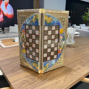 Puede incluir: Un juego de ajedrez decorativo con un marco dorado y obras de arte intrincadas. El juego presenta un diseño de tablero de ajedrez detallado con un borde dorado y un diseño circular azul y dorado. Las piezas de ajedrez no son visibles.