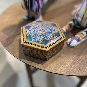May include: An ornate, hexagonal wooden box with a detailed, colorful design. The lid features a blue background with a central floral pattern and intricate gold and red accents. The box's sides have a geometric pattern.
