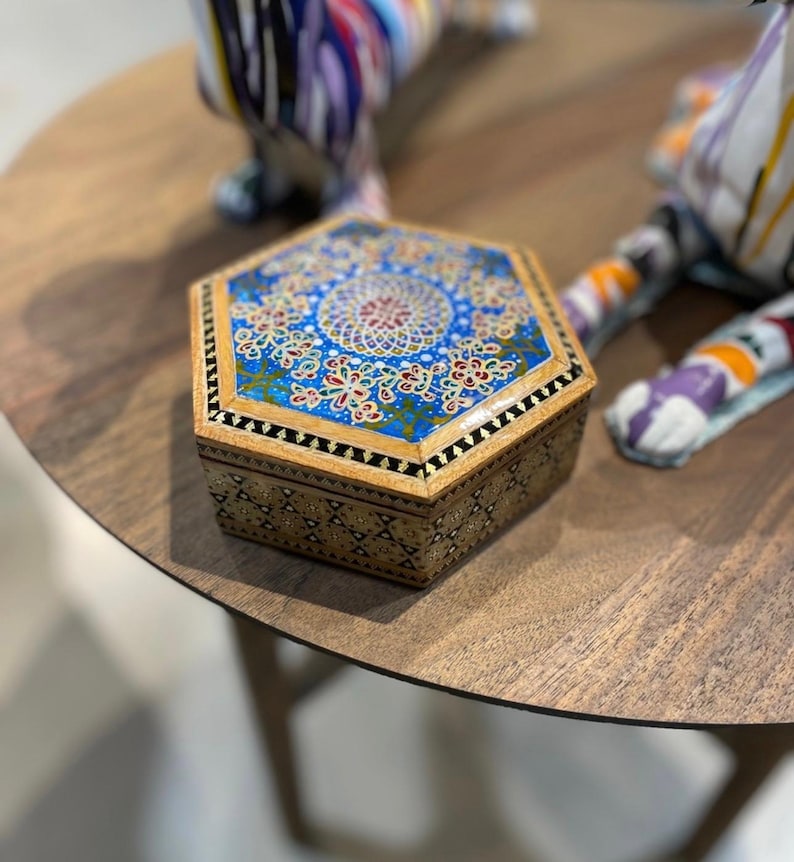 May include: An ornate, hexagonal wooden box with a detailed, colorful design. The lid features a blue background with a central floral pattern and intricate gold and red accents. The box's sides have a geometric pattern.