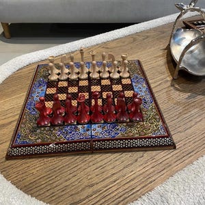 May include: A decorative chess set with a wooden board featuring a colorful, patterned border. The chess pieces are red and natural wood. The board is set up, ready for a game.