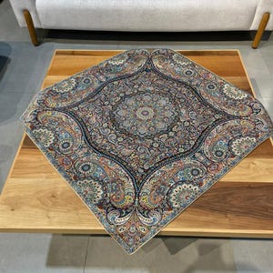 May include: A square, intricately patterned rug with a central medallion design. The rug features a complex floral and paisley pattern in shades of blue, orange, and cream. It is placed on a wooden coffee table, with a light-colored sofa in the background.