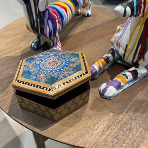 May include: An open, hexagonal wooden jewelry box with a detailed, colorful floral design on the lid. The box is on a wooden table, with two decorative dachshund sculptures nearby. The sculptures are painted with bright, abstract patterns.