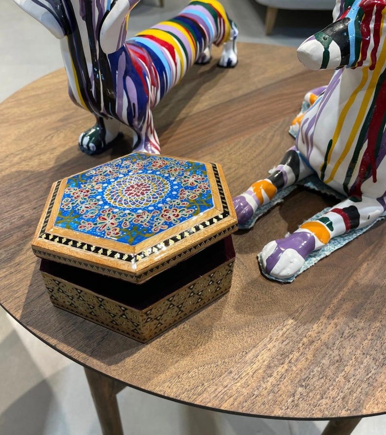 May include: An open, hexagonal wooden jewelry box with a detailed, colorful floral design on the lid. The box is on a wooden table, with two decorative dachshund sculptures nearby. The sculptures are painted with bright, abstract patterns.
