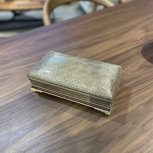 Puede incluir: Caja decorativa rectangular con tapa estampada en tonos dorados, beige y marrón. La caja tiene un borde marrón oscuro con una fina raya blanca y descansa sobre patas blanquecinas. La caja está sobre una superficie de madera.