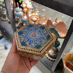 May include: A handcrafted, hexagonal wooden box with a detailed, colorful mosaic design on the lid. The mosaic features a blue background with floral patterns in red, gold, and white. The box is held in a hand.