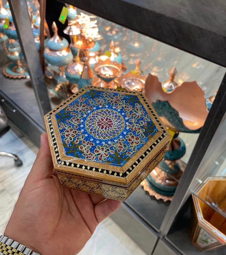 May include: A handcrafted, hexagonal wooden box with a detailed, colorful mosaic design on the lid. The mosaic features a blue background with floral patterns in red, gold, and white. The box is held in a hand.