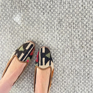 Puede incluir: Un par de zapatos sin cordones estampados con ribete marrón. Los zapatos presentan un diseño geométrico en negro, verde, rojo y beige. Los zapatos están sobre una superficie gris texturizada.