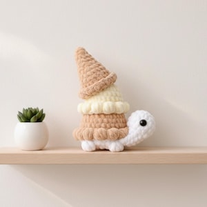 May include: A handmade crochet turtle with an ice cream cone design. The turtle has a white head and feet, with a tan and cream-coloured cone. A small succulent in a white pot sits on a wooden shelf nearby.