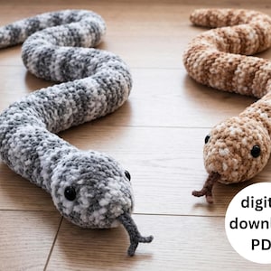 May include: Two plush crochet snakes are shown. One is grey and white, the other is brown and tan. Both have black eyes and a forked tongue. The snakes are on a wooden surface. The image includes a digital download PDF graphic.