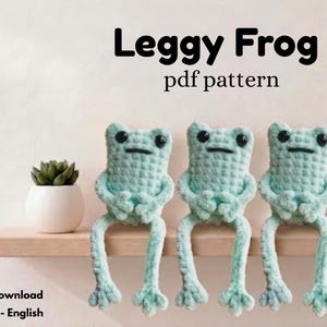 May include: Three light green crocheted frogs with long legs and black eyes sit in a row. The text Leggy Frog pdf pattern is displayed above the frogs, and the text PDF Digital Download US-Term - English is visible in the bottom left corner.