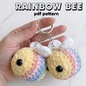 May include: Two rainbow-colored bee keychains with black eyes and white wings are held by a hand. The keychains are attached to a silver keyring. The text "RAINBOW BEE pdf pattern" is at the top.