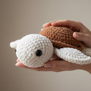 May include: A handmade crochet turtle in shades of brown and white. The turtle has a textured shell and head, with black button eyes. The turtle is being held in someone's hands.