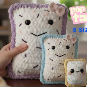 May include: Crocheted Pop-Tart plushies in three sizes. Each plush has a white base with black eyes, a stitched smile, and sprinkles. The largest has a purple border, the middle one has a blue border, and the smallest has a yellow border. Each has a bow.