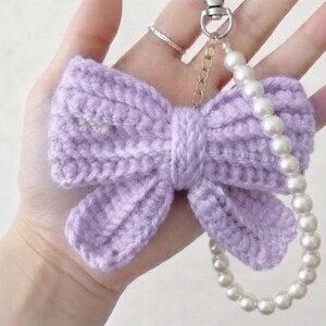 Può includere: Un portachiavi a forma di fiocco all'uncinetto color viola chiaro con una struttura in filato testurizzato. Il fiocco è fissato a un moschettone in metallo argentato e a una catenella, decorata con un filo di perle bianche finte a cerchio.
